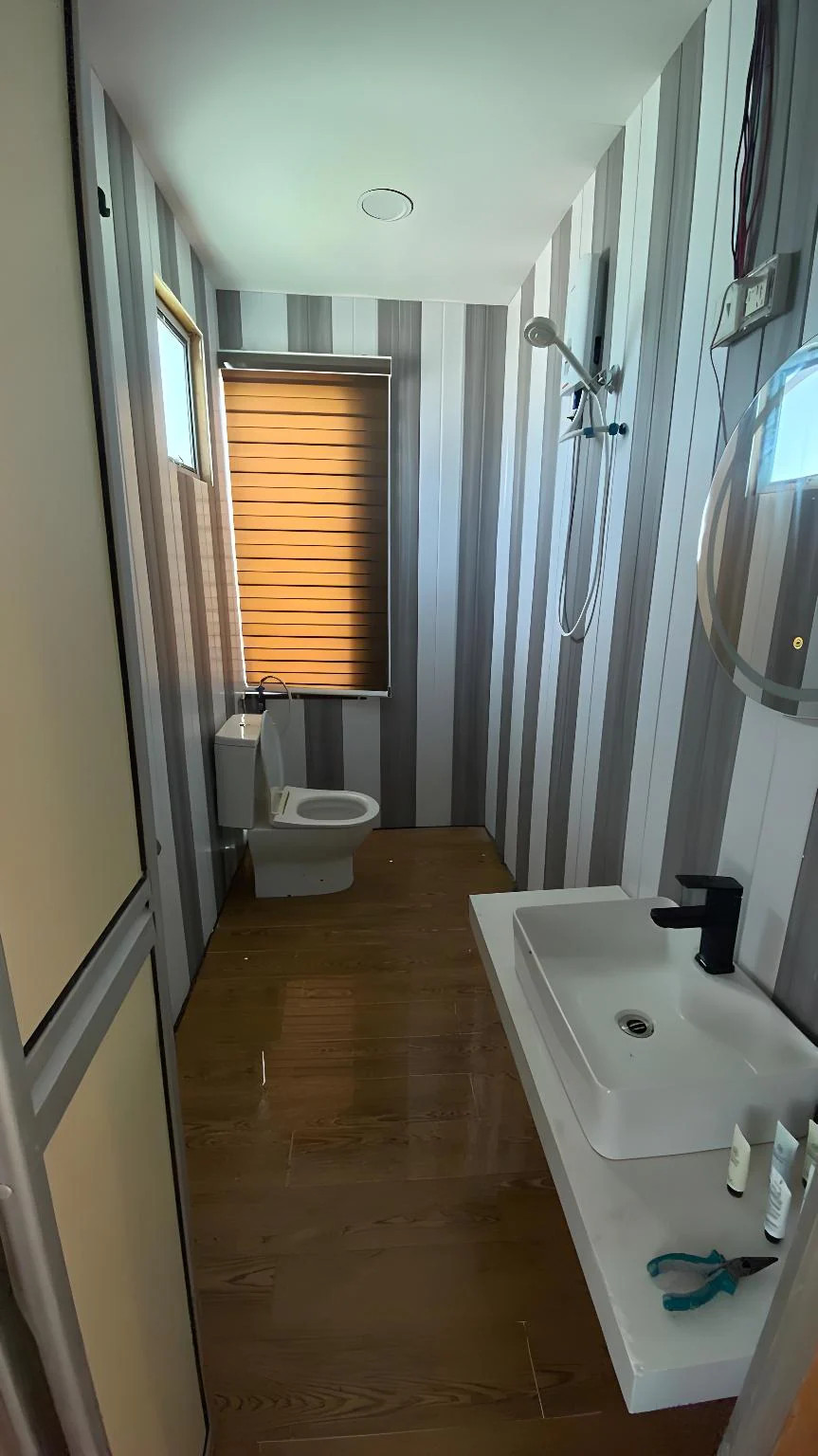 A modern bathroom at Blue Ocean Resort showcasing a clean white washbasin, a large vanity mirror, a ceramic toilet, tiled walls, and a glass-partitioned electric shower area.