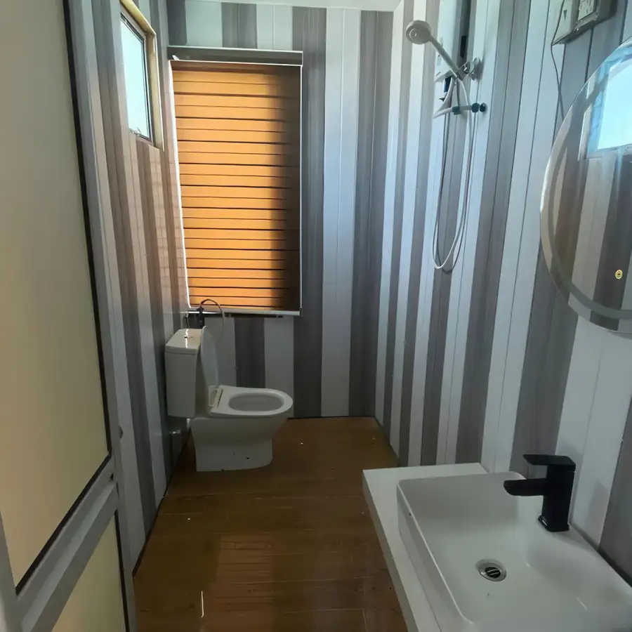 A modern bathroom at Blue Ocean Resort showcasing a clean white washbasin, a large vanity mirror, a ceramic toilet, tiled walls, and a glass-partitioned electric shower area.