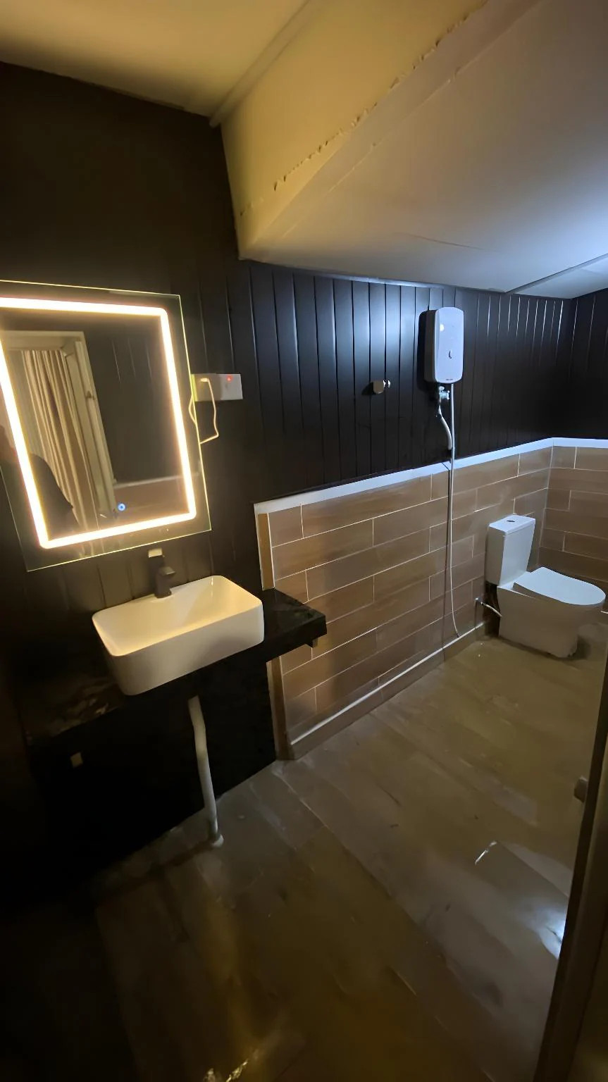 A spacious family bathroom at Blue Ocean Resort offering a well-lit vanity sink, a wall mirror, a white toilet, neutral floor tiles, and a dedicated glass electric shower enclosure.