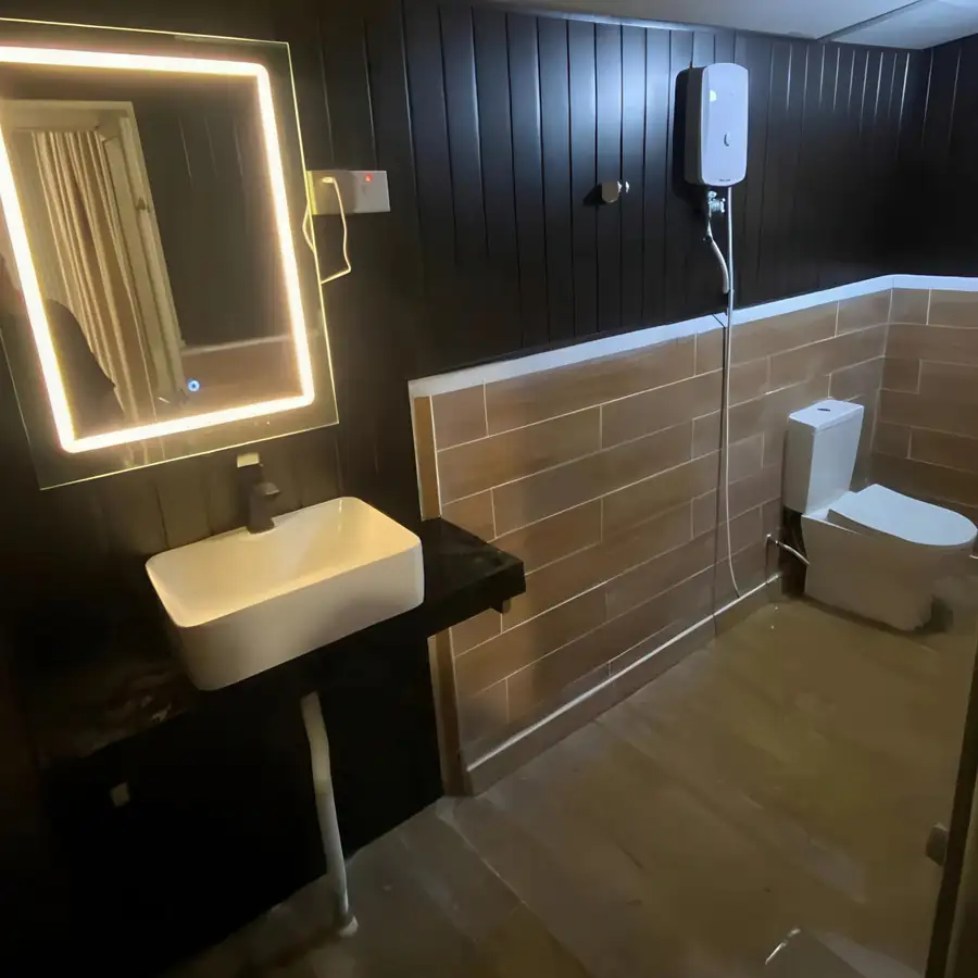 A spacious family bathroom at Blue Ocean Resort offering a well-lit vanity sink, a wall mirror, a white toilet, neutral floor tiles, and a dedicated glass electric shower enclosure.